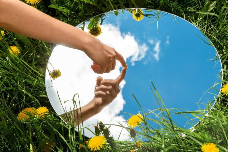 A hand reaches toward its reflection in a circular mirror lying in a grassy field with dandelions and a blue sky above.